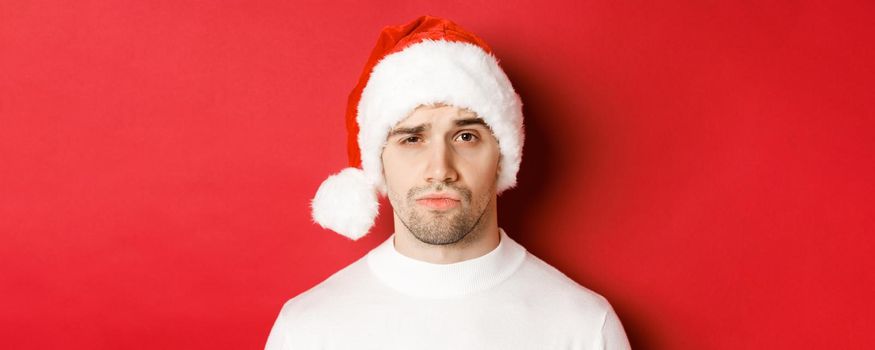 Close-up of doubtful frowning man, wearing santa hat and looking at camera uncertain, standing against red background.