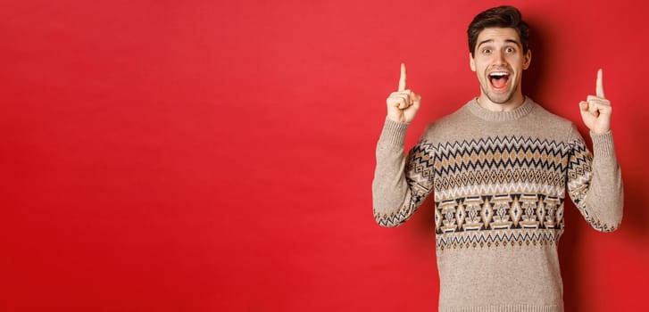 Image of handsome excited man, feeling happy about christmas holidays, pointing fingers up and looking cheerful, showing advertisement, standing in sweater over red background.