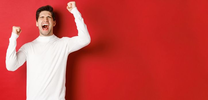 Portrait of handsome man in white sweater, feeling cheerful, celebrating victory, shouting for joy and raising hands up in victory, standing over red background.