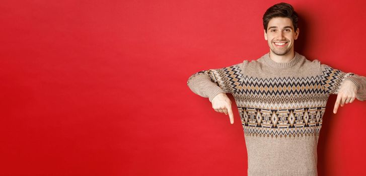 Concept of christmas celebration, winter holidays and lifestyle. Attractive happy man in xmas sweater showing promotion, pointing fingers down at logo, standing over red background.
