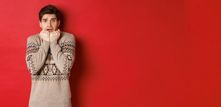 Portrait of scared and timid man in christmas sweater, looking frightened, watching something scary on new year, standing over red background.