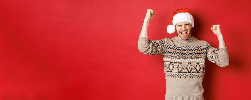 Satisfied handsome man, feeling lucky and happy, shouting for joy and making fist pumps, celebrating victory or win, receive awesome christmas gift, standing in santa hat over red background.