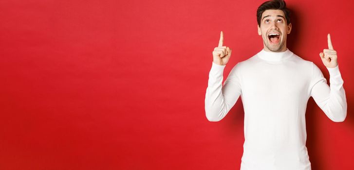 Concept of winter holidays. Image of amazed handsome guy reacting to christmas announcement, pointing and looking up at copy space, standing over red background.