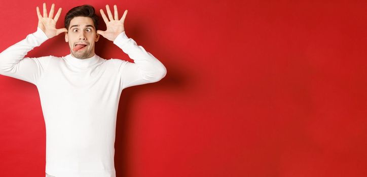 Portrait of funny caucasian guy, showing tongue and making faces, wearing white sweater, standing against red background.