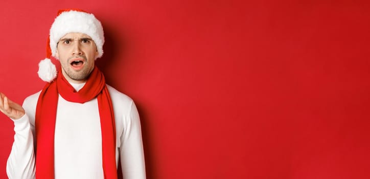Concept of christmas, winter holidays and celebration. Portrait of confused man in santa hat and scarf, frowning and looking perplexed, standing over red background.