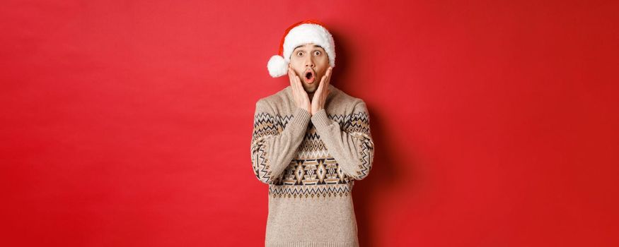Image of amazed and speechless man in santa hat, drop jaw and staring at awesome christmas offer, standing over red background.