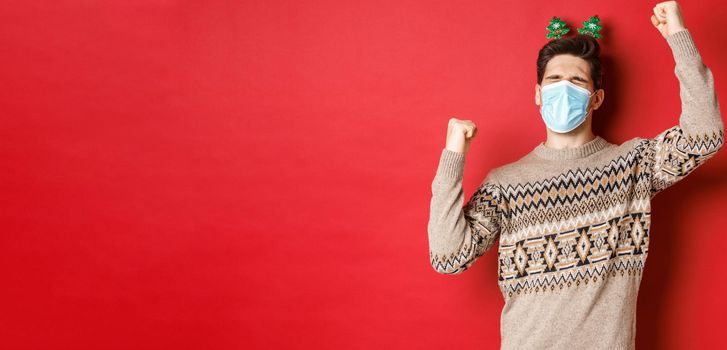 Concept of coronavirus, winter holidays and social distancing. Image of excited and happy man in medical mask and christmas clothing, raising hands up and celebrating new year, red background.