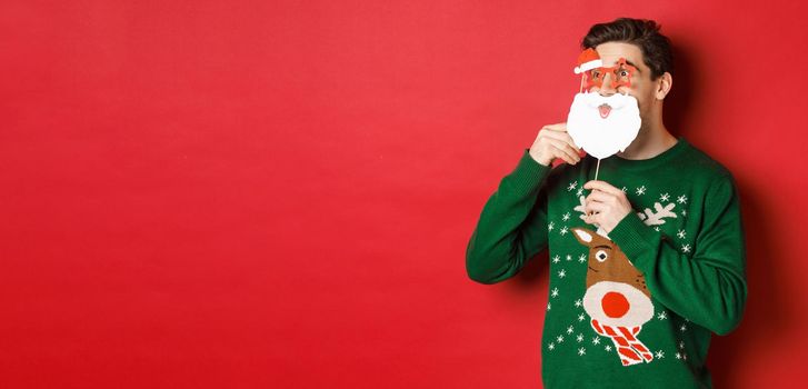 Portrait of surprised man in green christmas sweater, holding funny santa claus mask, celebrating new year, looking left amazed, sanding over red background.