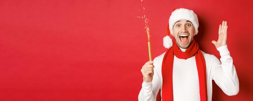 Concept of christmas, winter holidays and celebration. Handsome man celebrating new year and having fun, holding sparkler and smiling, wearing santa hat, standing over red background.