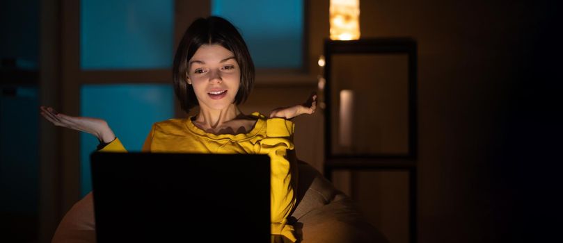 Portrait of beautiful caucasian girl uses laptop while sitting at home. In the evening creative woman works on a computer in her cozy living room.