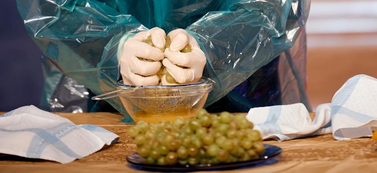 man with his gloves in a raincoat crushes grapes into a glass plate . High quality photo