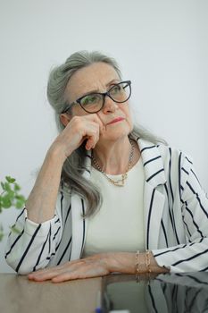 Tired senior grey haired businesswoman in striped jacket with eyeglasses is working in her office sitting at the desk and feeling bad due to menopause, menopause relief concept.