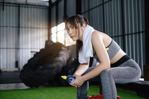 Woman wearing sport sitting and resting after exercise in fitness gym holding drinking water. Healthy people concept.