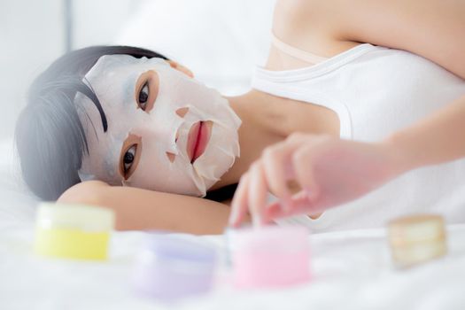 Beautiful young asian woman with sheet facial mask on bed at bedroom, beauty girl lying applying cosmetic and makeup with skin care face for wrinkle at home, one person, skincare treatment and health.