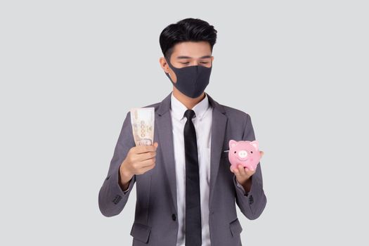 Young asian businessman in face mask holding money Thai and piggy bank isolated on white background, crisis of economic, business man in mask investment finance and planning saving during covid-19.