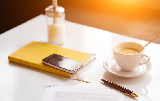 on the white table yellow notepad phone cup coffee pen and sheet of paper with a blurry dough . High quality photo
