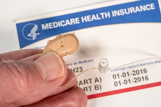 Macro close up of fingers holding tiny modern hearing aid on medicare card background as healthcare coverage is improved
