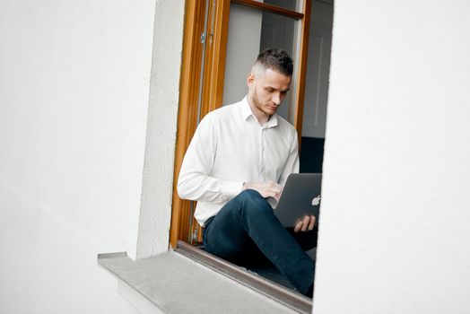 a young guy sits on the windowsill at home and works with a laptop. High quality photo