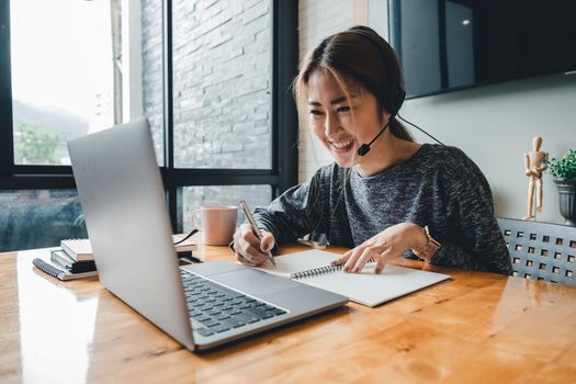 Happy asian woman student watching lesson online and studying from home. Young woman taking notes while looking at computer screen following professor doing math on video call.