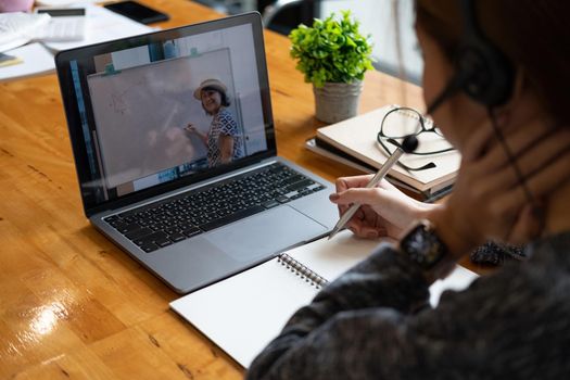 Young student watching lesson online and studying from home. Young woman taking notes while looking at computer screen following professor doing math on video call. Girl studying from home on laptop.