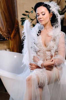 the bride, dressed in a boudoir transparent dress and underwear, sits on a vintage bathtub with a feather in her hands, The morning of the bride.