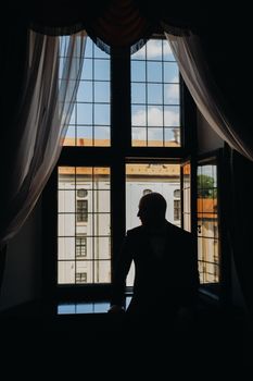 Portrait of the groom in a suit and bow tie sitting in the interior near the window.