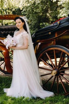 A stylish bride with a bouquet stands near a carriage in nature in retro style.