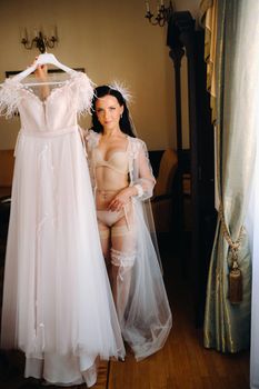 a bride dressed in a boudoir transparent dress and underwear holds her wedding dress in her hands in the interior of the house.