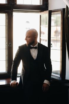 Portrait of the groom in a suit and bow tie sitting in the interior near the window.