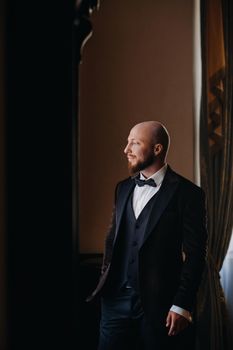 Portrait of the groom in a suit and bow tie, standing in the interior.