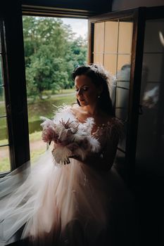 A bride in a wedding dress and a bouquet sits at an open old window and looks.