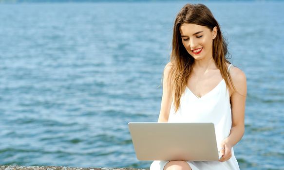 Smiling girl with laptop on her lap sits on a dock not the shore of the ocean lake. High quality photo