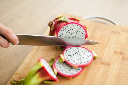 Female hands is cutting a dragon fruit or pitaya with pink skin and white pulp with black seeds on wooden cut board on the table. Exotic fruits, healthy eating concept.