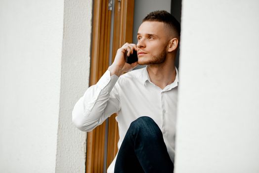 a serious man sits in the window opening of the house and talks on the phone. High quality photo