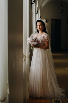 The bride in a wedding dress and with a bouquet stands at the old window and looks.