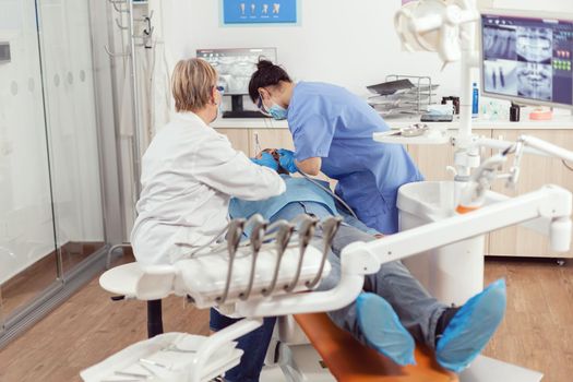 Stomatologist senior woman doing hygienic cleaning of teeth using sterile dental tools. Patient sitting with open mouth on dental chair, dentist doctor performing examination in modern clinic