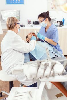 Stomatologist senior woman performing examination and taking care of teeth using dental tools. Orthodontist speaking to sick man sitting on stomatological chair in modern clinic