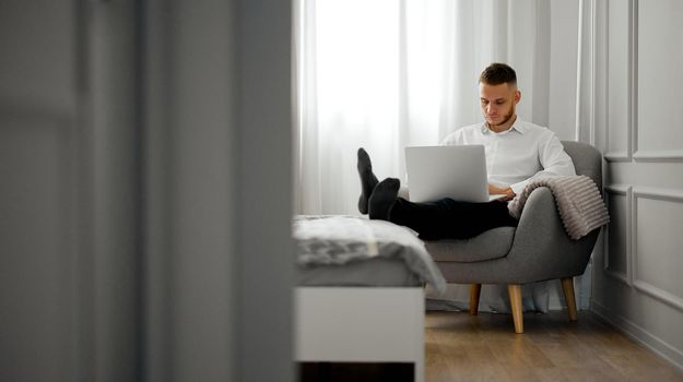 A young man in a shirt works at a laptop in the bedroom on a chair with legs on the bed. High quality photo