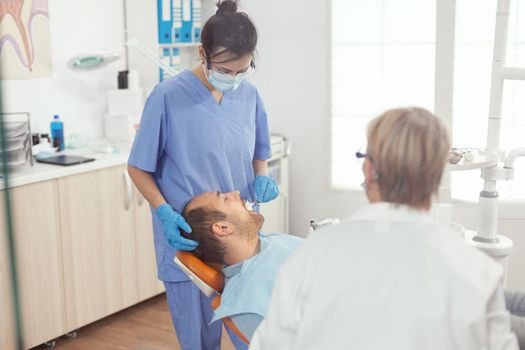 Medical nurse making professional teeth cleaning to man patient during orthodontic consultation in stomatology office. Hospital team examining toothache preparing tooth treatment