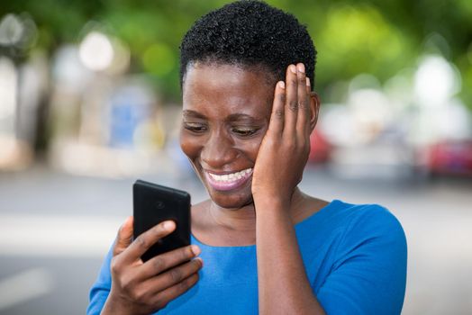 young girl standing in blue camisole looking at mobile phone laughing with hand on cheek.