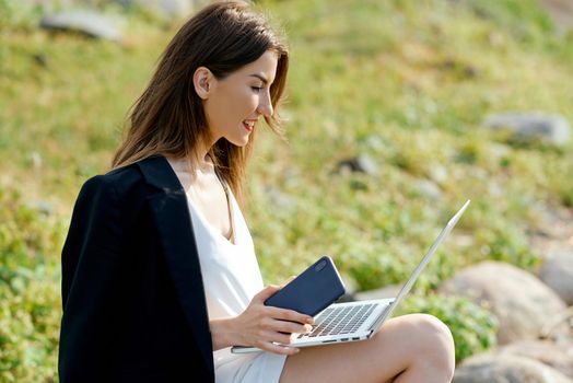 Businesswoman on a stone on the beach works behind a laptop in a white dress and black jacket . High quality photo