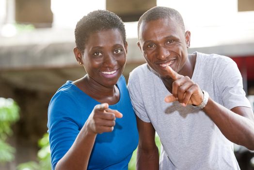 young african couple standing pointing at each other and looking at camera laughing.
