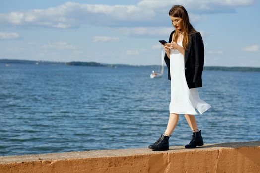 Woman with phone in hand walks across pier in white dress and jacket stares at screen. High quality photo