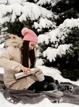 Merry Christmas and happy New Year. Beautiful woman in warm winter clothes holding thermos and drinking hot tea or coffee outdoors in snowy day