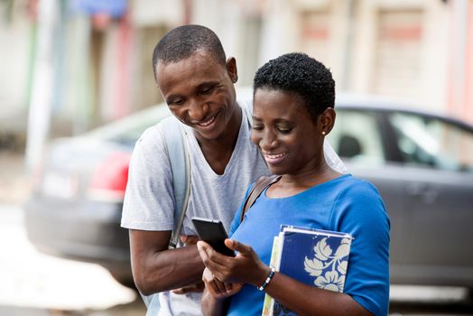 Two young happy college students standing in the street are watching and reading a message on a smart phone outdoors.