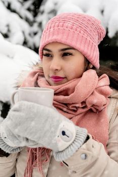 Merry Christmas and happy New Year. Beautiful woman in warm winter clothes holding mug drinking hot tea or coffee outdoors in snowy day