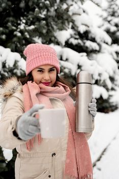 Merry Christmas and happy New Year. Beautiful woman in warm winter clothes holding thermos and drinking hot tea or coffee outdoors in snowy day