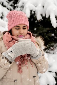 Merry Christmas and happy New Year. Beautiful woman in warm winter clothes holding mug drinking hot tea or coffee outdoors in snowy day