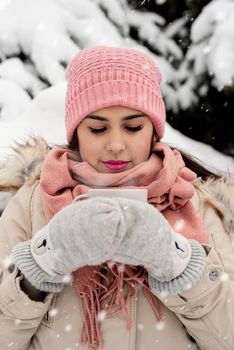 Merry Christmas and happy New Year. Beautiful woman in warm winter clothes holding mug drinking hot tea or coffee outdoors in snowy day