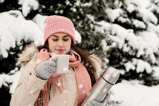 Merry Christmas and happy New Year. Beautiful woman in warm winter clothes holding thermos and drinking hot tea or coffee outdoors in snowy day